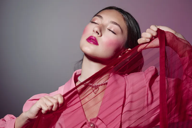 Young woman with glossy pink lips and vibrant blush poses holding a red sheer fabric against grey studio background. Concept for creative beauty, fashion, skin care, and editorial photography.