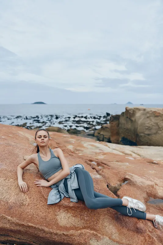 Young athletic woman in gray sportswear poses on rocky beach, evoking serenity and confidence with ocean in background, perfect for fitness and wellness themes