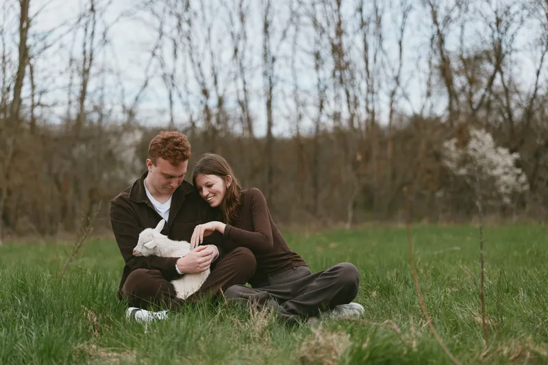Couple outdoors with a baby goat in a grassy field, sharing warm smiles and tender moments, illustrating a cozy family lifestyle and harmony with nature in a peaceful rural setting