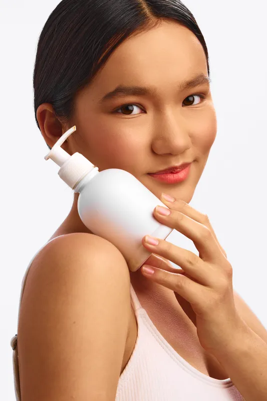 Asian woman is shown holding a cosmetic bottle near her cheek in a clean studio setting. She smiles softly, presenting a calm and confident skincare moment.