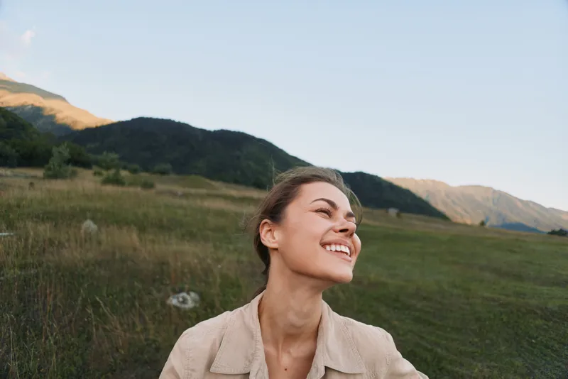 A woman in a light jacket smiles in a sunlit meadow, with rolling hills in the background, capturing a candid outdoor lifestyle moment of happiness.