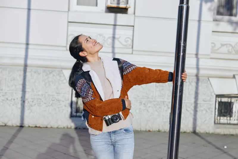 Smiling woman outdoor street pole casual fashion joy urban pose in daylight candid moment set against a street backdrop, jacket and jeans, lifestyle photography capturing a lively, fashionable moment