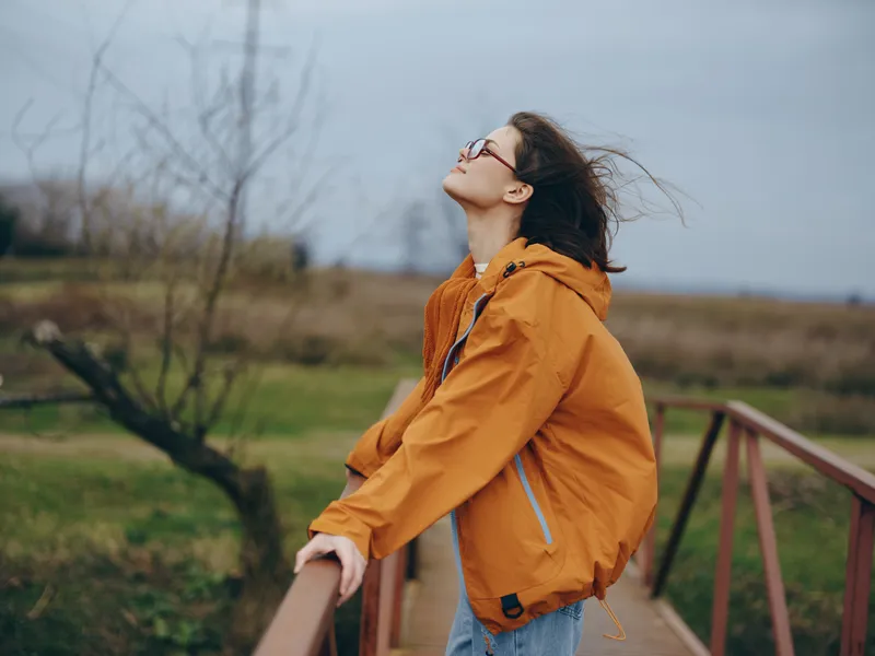 Woman lifestyle outdoors orange jacket, wooden railing, wind blowing hair, smiling while looking up, soft daylight, countryside meadow background, relaxed standing posture