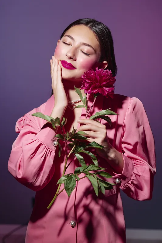 Young woman in a pink long-sleeve dress holds a large dark pink flower against a purple background. Her eyes are closed, lips shiny with deep lipstick. Concept of beauty and fashion.