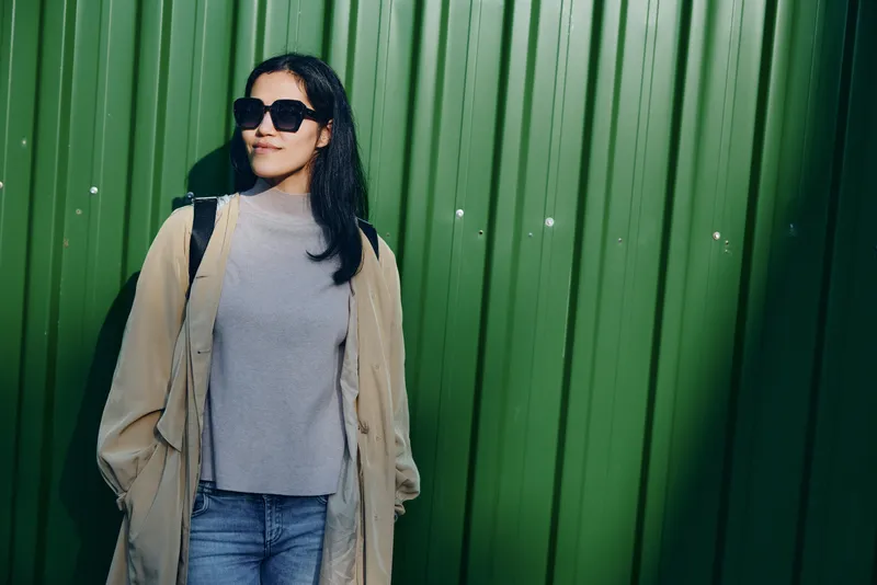 Woman in sunglasses leans against a green wall, wearing a beige coat over a gray knit top and jeans, creating a casual street style scene with urban attitude and warm lighting.