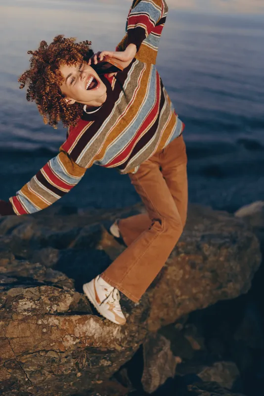 Woman lifestyle ocean portrait with curly hair, striped sweater, and tan pants, posing on rocky coast at golden hour, relaxed expression, casual outdoor fashion in sea air today
