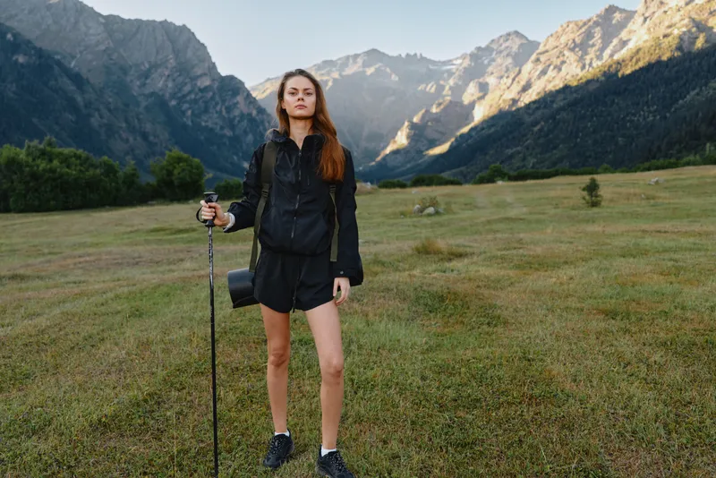 A lone female hiker stands in a grassy valley with a trekking pole, set against dramatic mountains. She wears a dark outfit and looks ready for outdoor adventure.