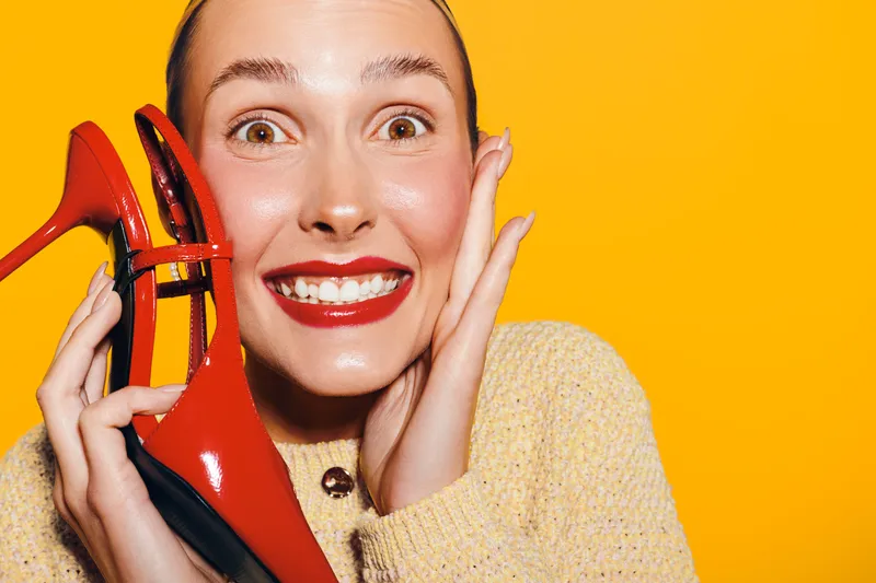 Happy woman with bright red lipstick holds red high heel shoe near face, smiling widely on yellow background. Concept of fashion, style, beauty, and joyful emotions.