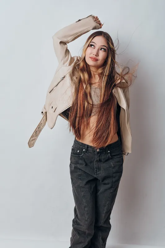 Young woman with long hair in casual outfit posing with raised arm and looking up on white background. Stylish female model wearing jacket and jeans for fashion or lifestyle.