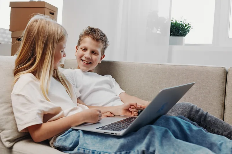 Two smiling children sitting on a couch using laptop together in a cozy living room with natural light. Concept of technology, childhood, education, and friendship.