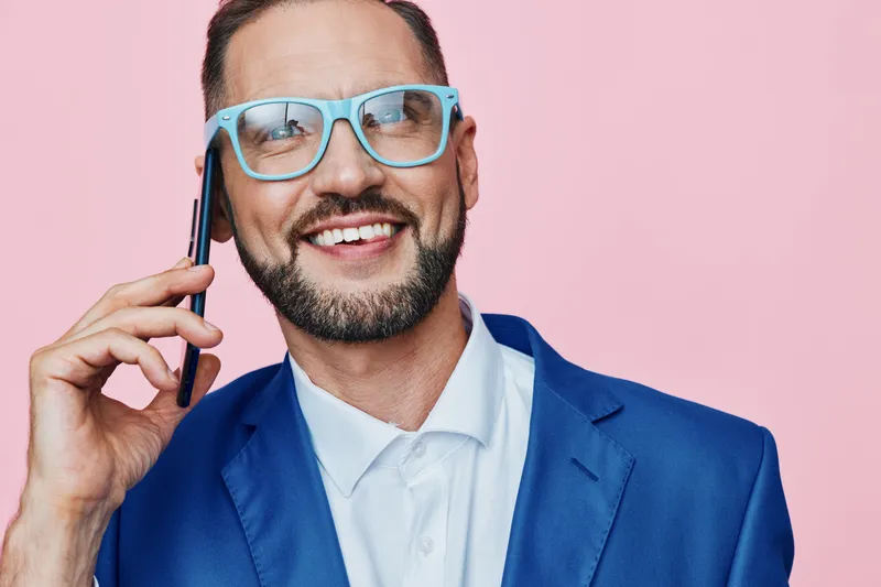 A professional business man on a pink isolated background, wearing a blue suit and glasses, talking on a phone with a confident smile, conveying focus, energy, and approachable leadership.