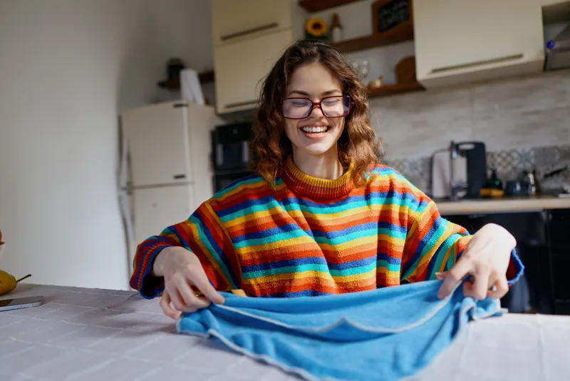 Woman at home irons clothes with a smile, wearing a colorful striped sweater, in a bright kitchen, creating a cheerful casual laundry moment indoors