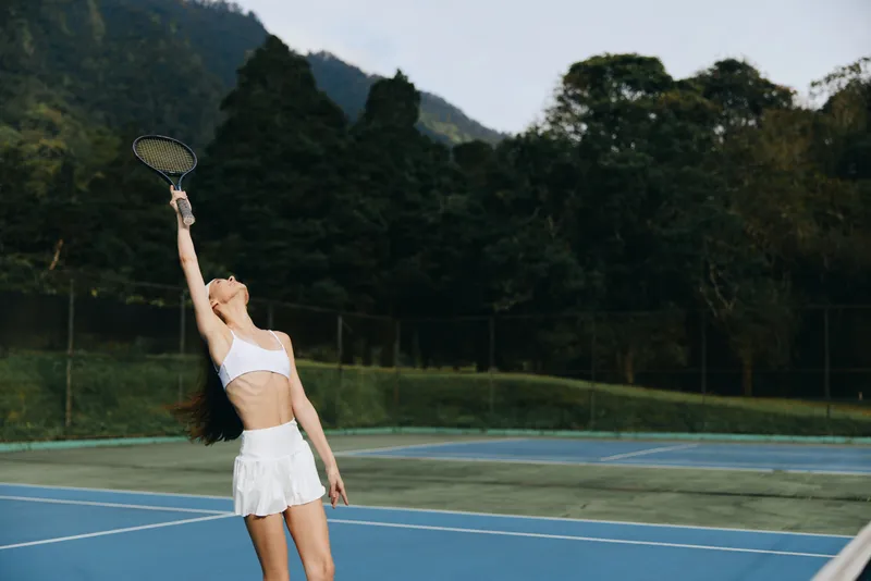 Athletic young woman playing tennis outdoors, expressing joy, wearing a white sporty outfit, on a green tennis court with a vibrant background