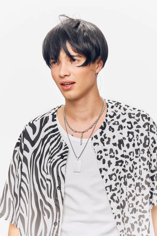 young man with patterned shirt and necklace looks curious against white background, wearing black short hair and casual style in studio portrait photography.