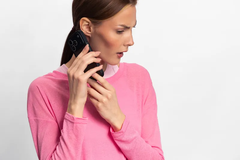 Surprised young woman in pink sweater holds smartphone near ear, looking aside. Isolated on white background. Concept of unexpected phone call, communication, and reaction.
