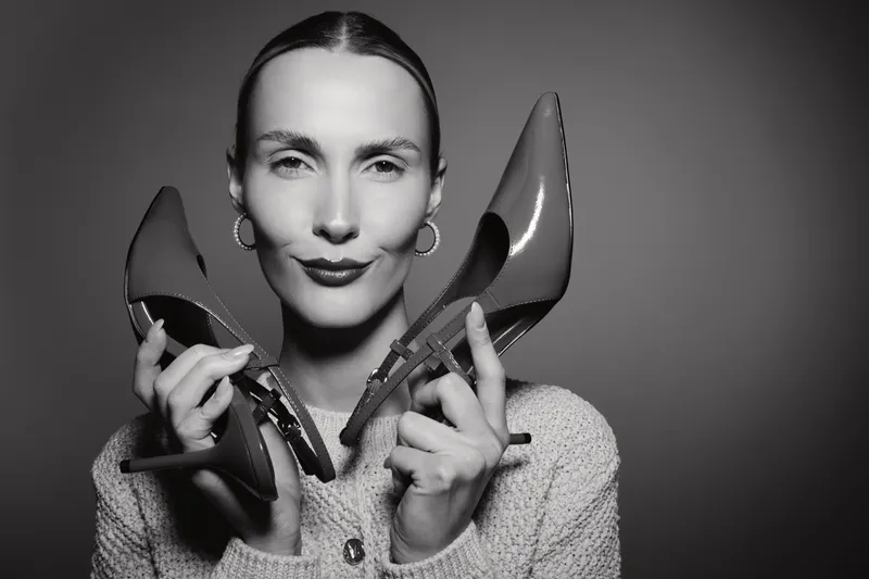 Young white woman with light skin holds high heel shoes near face, wearing sweater and earrings, posing on grey background in studio.