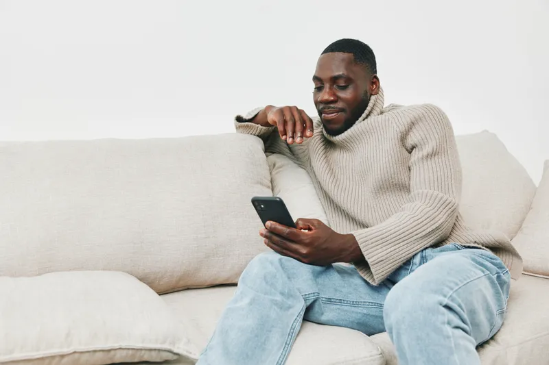 Young African man relaxing on a couch, using a smartphone while wearing a cozy sweater and jeans, showcasing a casual, modern lifestyle
