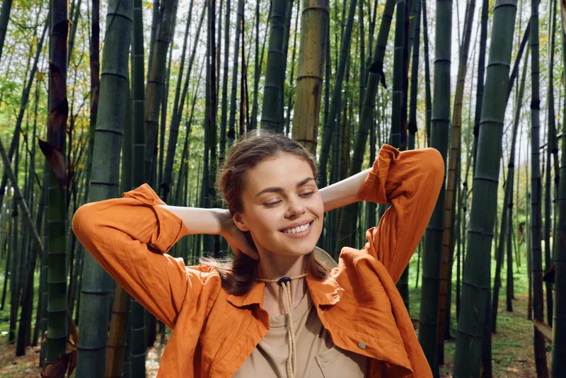 woman, smile, bamboo, forest, nature, relaxation Young woman with eyes closed smiling in a bamboo forest wearing orange jacket, enjoying serene sunlight, calm posture and peaceful outdoor wellbeing.