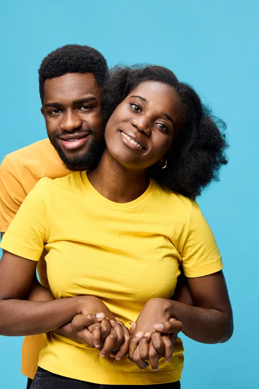 Young African couple smiling together in matching yellow t shirts against a blue background, showcasing love and happiness in a vibrant, joyful setting
