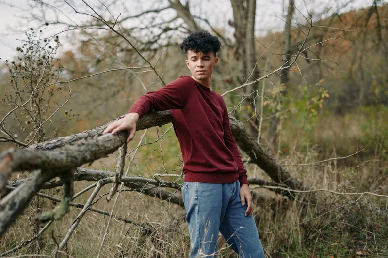 Authentic portrait of a young man leaning on a rustic fence in a natural field, capturing genuine posture and quiet confidence in an everyday outdoor setting.