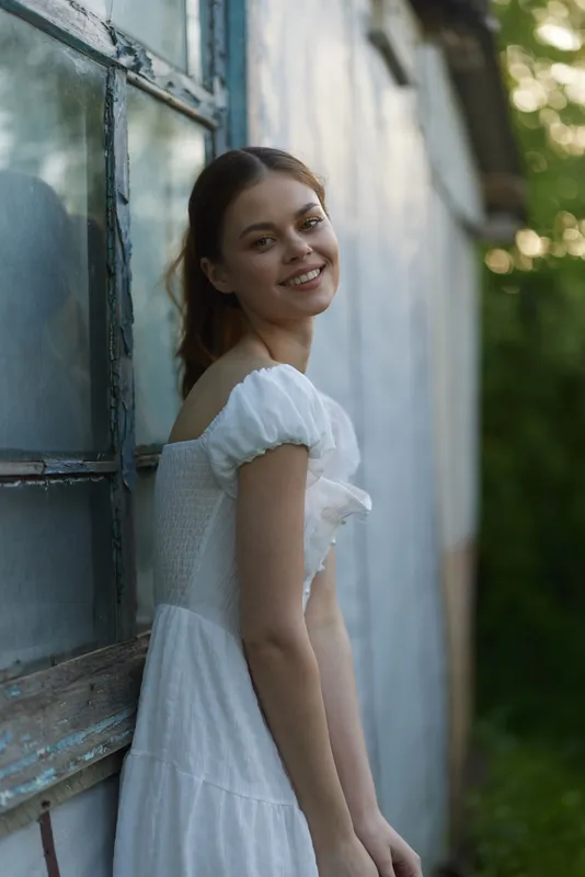A smiling young woman of Eastern European descent in a white summer dress stands beside an old building, capturing the essence of joy and warmth in nature