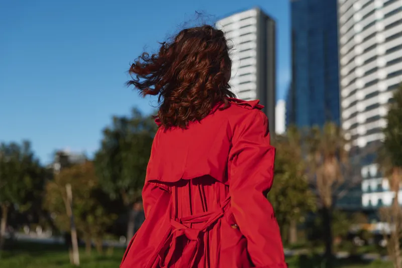 Redcoat woman stands outdoors with back to camera, wind in hair, modern city skyline behind, vibrant urban mood and confident style with energy and flair