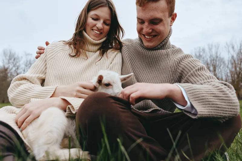 Cheerful couple outdoors, gently patting a curious goat kid, knit sweaters, sunlit grass, calm atmosphere, natural colors, candid moment, joyful connection, rural setting