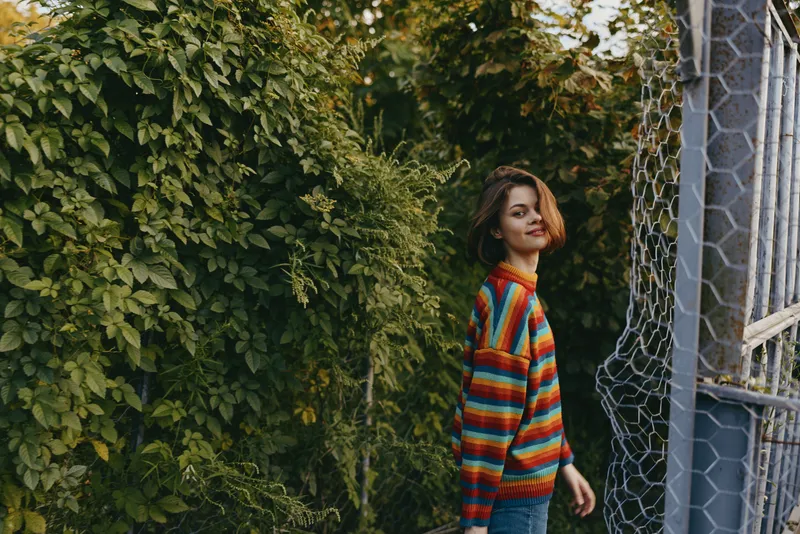 Woman in a colorful striped sweater smiling with short hair against green foliage and chainlink fence, casual outfit in an outdoor portrait of a young adult walking and turning back.