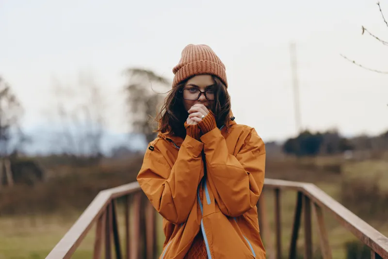 Woman lifestyle outdoors bridge winter beanie scarf orange coat, standing on wooden bridge, hands near mouth, enjoying cold fresh air, soft daylight, background blurred countryside, cozy calm mood