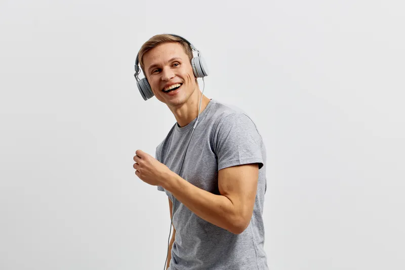 Smiling young man wearing casual gray t-shirt and large headphones, enjoying music with joyful expression on plain white background, studio portrait. People lifestyle concept
