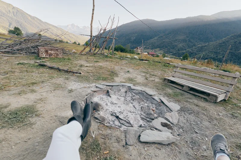 Campfire mountains boots bench firepit hiking outdoors relaxation. Two people resting with boots stretched toward a stone firepit and wooden bench, panoramic mountain valley view at campsite.