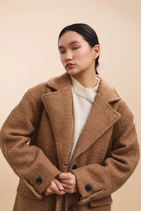 An asian woman wearing a warm brown coat stands in a soft studio backdrop, exuding calm confidence and refined style in a minimalist fashion portrait.