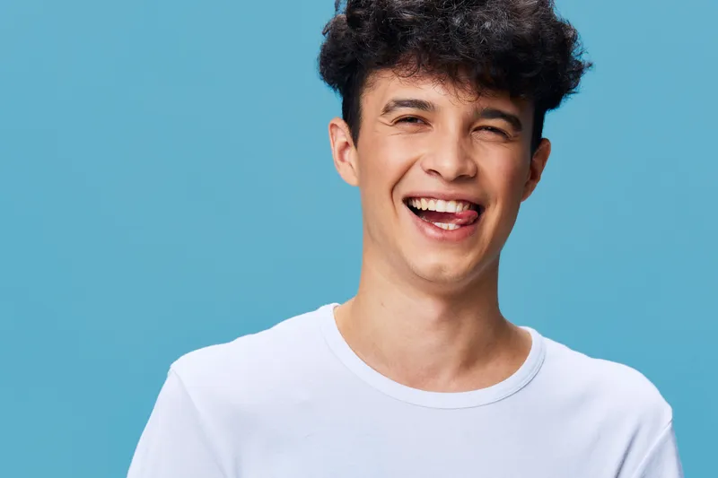 Smiling young man with curly hair wearing white t-shirt against a blue background, expressing joy and happiness, casual style portrait, studio lighting.