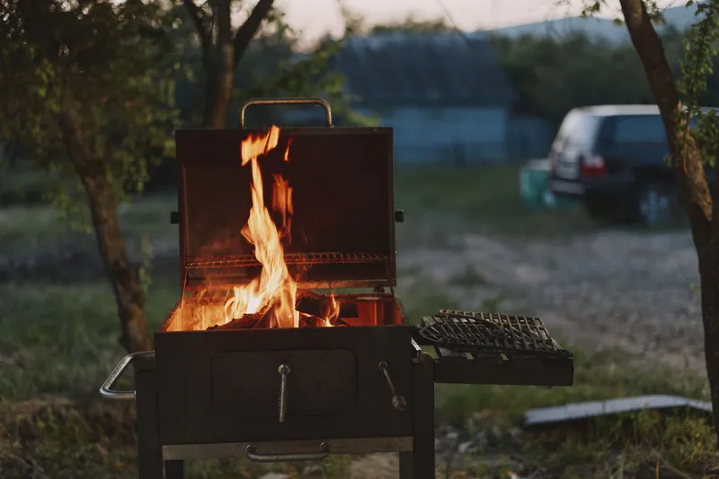 Outdoor barbecue grill with vibrant flames, set in a grassy yard at sunset, creating a warm, inviting atmosphere for gatherings and summer celebrations