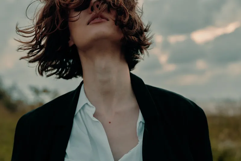 Fashion in a windy outdoor setting, a blazer and open shirt under a moody sky, close cropping emphasizes movement, confidence, and bold style in natural light.