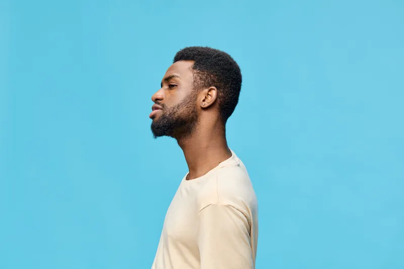 Profile portrait of a young Black man with a thoughtful expression against a bright blue background, showcasing modern casual fashion and seasonal style