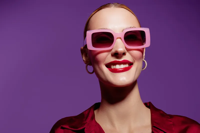 Smiling woman with red lipstick wearing pink sunglasses and hoop earrings poses against purple background. Fashion portrait with confident expression and stylish accessories in studio lighting.