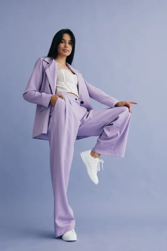 Stylish woman in lilac pantsuit poses confidently against a purple backdrop, wearing white sneakers and a lace top, showcasing modern business fashion and elegance.