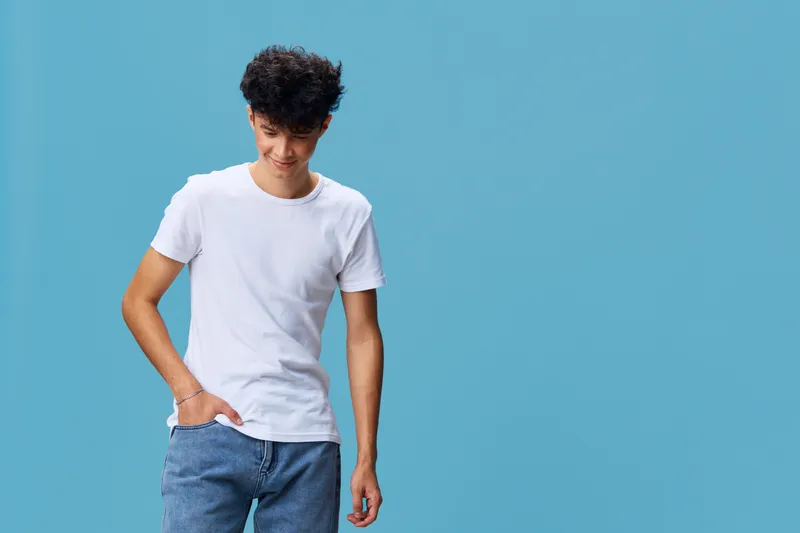 smiling young man in casual white t-shirt and blue jeans standing against blue background, looking down with relaxed posture and natural curly hair
