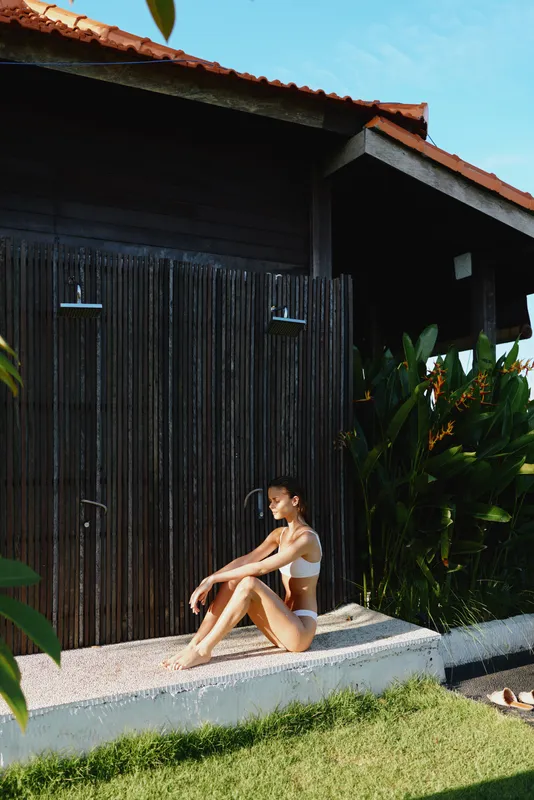 Woman in bikini sits on a concrete ledge beside a dark wooden fence, tropical plants framing the scene. Outdoor relaxation, summer lifestyle, and sunlight convey calm, simple joy outdoors.