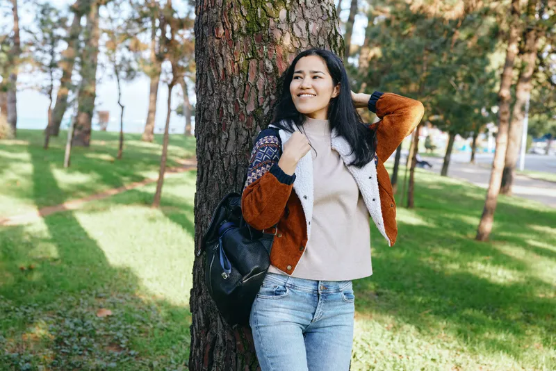 Woman leaning against a tree in a park, wearing a cozy jacket and sweater, smiling as she enjoys a sunny outdoor moment among green grass and tall pines