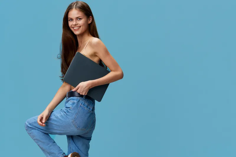 smiling woman holding laptop wearing casual jeans and black top posing confidently on blue background with long hair, youthful, bright, cheerful atmosphere