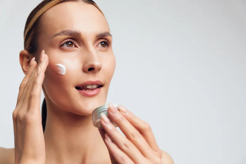 Young woman applies moisturizing cream for face skincare application, gentle routine, hands smoothing texture on cheek, spa close up beauty portrait, soft light, clean background, fresh glow