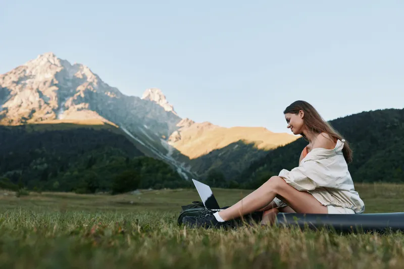 A relaxed woman sits on a grassy field with a laptop, enjoying nature and work balance in a peaceful outdoor office scene, framed by distant mountains and clear sky.