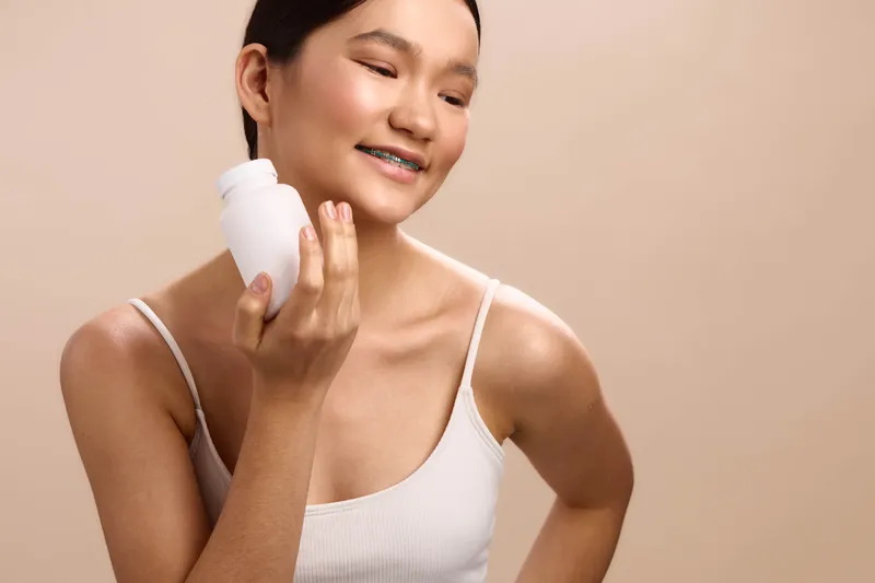 Asian woman skincare routine in a clean studio, applying lotion from a bottle with a gentle smile, presenting a calm and healthy beauty moment in a simple, modern setting.