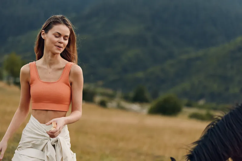 A smiling woman stands in a golden meadow beside a horse, wearing an orange crop top and a light jacket tied at the waist. She appears relaxed, enjoying outdoor life and sunny countryside moments.