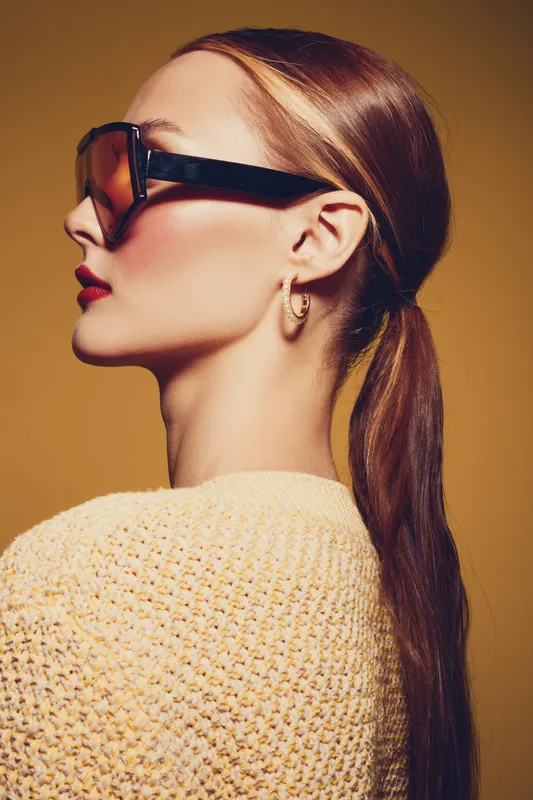 Profile of a stylish woman with long brown hair tied back wearing large sunglasses and gold hoop earrings. She wears a textured yellow sweater against a warm brown background.