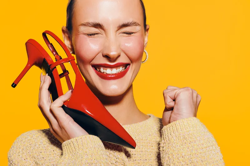 Happy woman with red high heels smiles with closed eyes on yellow background. Concept of fashion, style, joy, and vibrant color contrast in studio portrait.