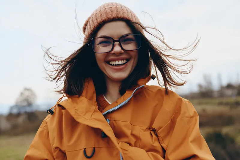 Woman outdoor lifestyle portrait, laughing in windy autumn, knit hat and orange coat, glasses, hair blowing, sunlit field background, candid moment, natural colors