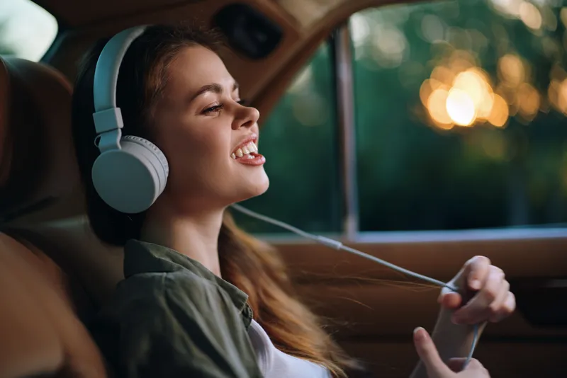 Young woman enjoying music with headphones in a car during sunset, reflecting happiness and relaxation in a cozy atmosphere.